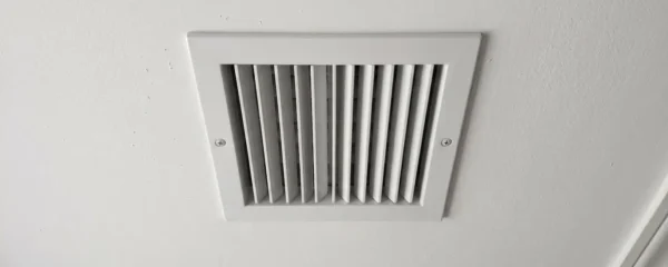 Propriétaire vu de dos observant une grille de ventilation au plafond dans un corridor résidentiel aux murs clairs