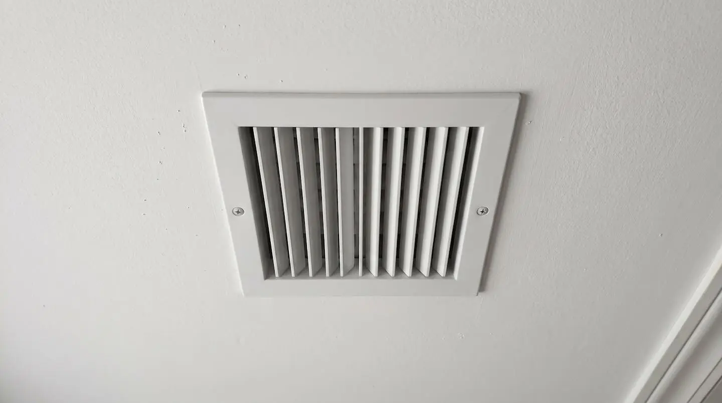 Propriétaire vu de dos observant une grille de ventilation au plafond dans un corridor résidentiel aux murs clairs